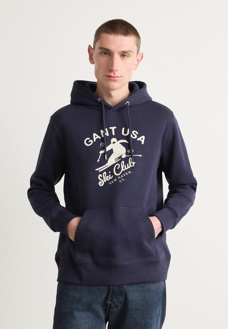 Sudadera azul marino de algodón con un bolsillo frontal, que presenta una impresión gráfica blanca de "GANT USA Ski Club" y acentos de diseño relacionados con el esquí.