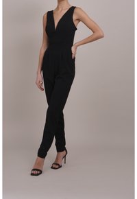 Mono negro sin mangas con escote en V, cintura ajustada, piernas cónicas y bolsillos laterales. Combinado con tacones altos negros con correas.