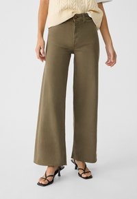 Olijfgroene wijde pantalon van denim, met een hoge taille en een rafelige zoom. Gecombineerd met een crème gebreide top en zwarte strap sandalen.