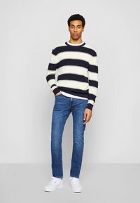 Modro-mornarsko pulover z kremasto črtami, narejen iz pletenega materiala, kombiniran z modrimi kavbojkami in belimi supergami. Preprost okrogel izrez, rebrasti manšeti in rob.