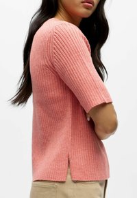 Roze ribgebreide trui met korte mouwen, voorzien van een gespleten zoom aan de zijkanten. De textuur oogt zacht en licht dik.