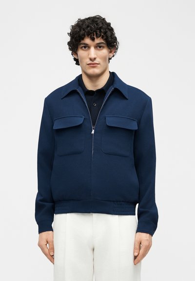 Νεαρός με σγουρά μαλλιά, ντυμένος με μπλε σκούρα ζακέτα με φερμουάρ και τσέπες με καπάκι, και άσπρο παντελόνι, στέκεται μπροστά σε ένα απλό φόντο.