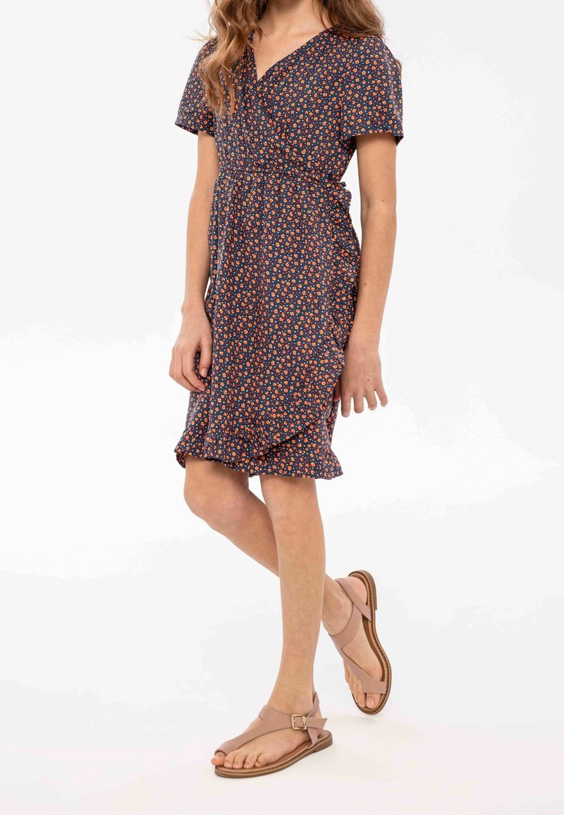 Femme portant une robe portefeuille fleurie bleu marine avec des volants et des sandales beiges, debout avec une jambe croisée devant l'autre.