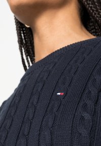 Pull en maille bleu marine avec motif torsadé et col côtelé. Il présente un petit logo en rouge, blanc et bleu sur l'épaule gauche.