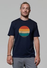Marineblauwe T-shirt met een ronde afbeelding die horizontale strepen in oranje, geel en teal kleuren toont, tegen een effen achtergrond.