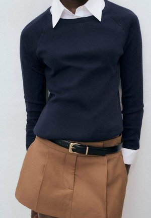 Personne portant un pull bleu marine par-dessus une chemise blanche à col, rentrée dans un pantalon marron taille haute avec une ceinture noire et une boucle dorée.