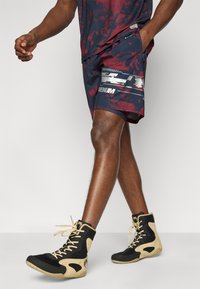 Αθλητικά σορτς με μοτίβο σε βουργουνδί και ναυτικό μπλε, με ασημένιες λεπτομέρειες, συνδυασμένα με αθλητικά παπούτσια ψηλού κορμού σε μαύρο και μπεζ. Έμφαση στην υφή και το σχέδιο.