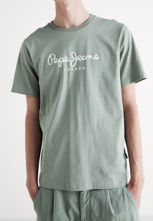 Henkilö pukeutunut vaaleanvihreään Pepe Jeans London -t-paitaan, jossa on lyhyet hihat, ja vastaaviin vihreisiin housuihin, kädet taskuissa.