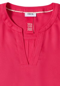 Blouse rose vif avec un col rond muni d'une petite fente, des manches courtes et un détail froncé à l'avant. Étiquettes visibles sur le col.
