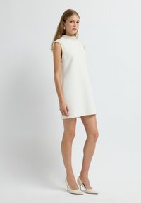 Witte mouwloze jurk met textuurstof, hoge hals en parelaccenten op de schouders. Gecombineerd met witte hoge hakken.