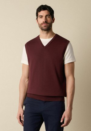 Mężczyzna ubrany w bordową kamizelkę z dekoltem w serek na białą koszulę z krótkim rękawem i ciemne spodnie, stojący na beżowym tle.