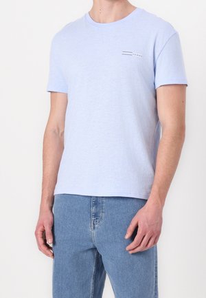 Homme portant un t-shirt bleu clair à manches courtes avec col rond et un jean bleu sur un fond blanc uni.