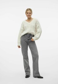 Un pull crème en maille torsadée avec un col en V, assorti à un jean gris évasé taille haute. Le modèle porte des bottines noires à la cheville et de petites boucles d'oreilles en créoles.