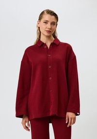 Camicia bordeaux con colletto, tessuto a costine, vestibilità oversize e maniche lunghe, con cinque bottoni lungo il davanti.