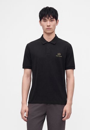 Giovane uomo che indossa una polo nera Armani Exchange a maniche corte e pantaloni grigi su sfondo bianco semplice.