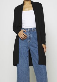 Cárdigan negro acanalado con mangas largas y apertura frontal. Combinado con jeans de mezclilla de tiro alto en azul claro, que presentan un cierre clásico de botón y cremallera.