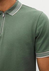 Vihreä poolopaita, joka on valmistettu teksturoidusta kankaasta ja jossa on vetoketjukaulus sekä raidalliset yksityiskohdat kauluksessa ja hihoissa.