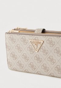 Portafoglio Guess beige con motivo a rilievo del logo, emblema triangolare Guess dorato e due cursori in metallo dorato su uno sfondo chiaro.