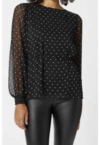 Blouse noire à pois avec manches longues transparentes, encolure ronde et poignets élastiques ; taille froncée ; texture en couches.