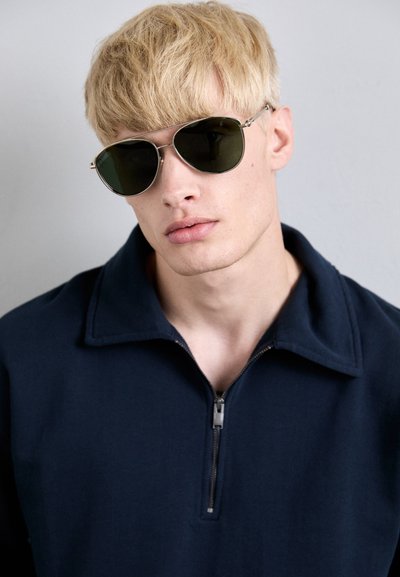 Navy zip-up collar jacket paired with silver-frame aviator sunglasses featuring green lenses; smooth texture, modern design, minimal detailing.