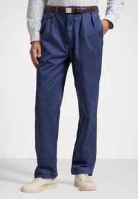 Pantalones de algodón azules con un corte relajado, que cuentan con pliegues frontales, bolsillos laterales y un cinturón de cuero marrón. Combinados con zapatillas de deporte en color beige claro.