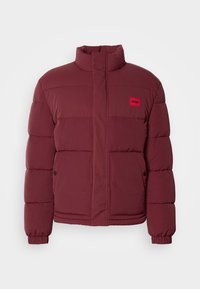 Bordeaux puffer-jakke med høj krave, quiltet tekstur og rød mærkepatch. Har trykknaplukning og sidelommer.