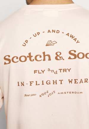 Dos d'un t-shirt beige avec texte marron incluant "Scotch & Soda," "Up - Up - And - Away,", une icône d'avion en papier, et "In-Flight Wear Amsterdam."