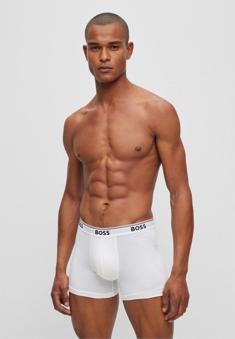 Weiße Herren-Boxershorts mit einem schwarzen Elastikbund, der mit "BOSS"-Branding versehen ist. Glatte Textur, körperbetontes Design und Länge bis zur Mitte des Oberschenkels.