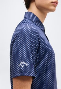 Άτομο που φοράει μπλε ναυτικό πουκάμισο πόλο με κοντά μανίκια και διακριτικό λευκό λογότυπο Callaway στο αριστερό μανίκι.