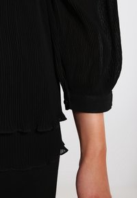 Czarna bluzka w plisowanym stylu z przezroczystym, teksturowanym materiałem, falbaniastym dołem i długimi rękawami z mankietami, nadająca warstwowy wygląd.