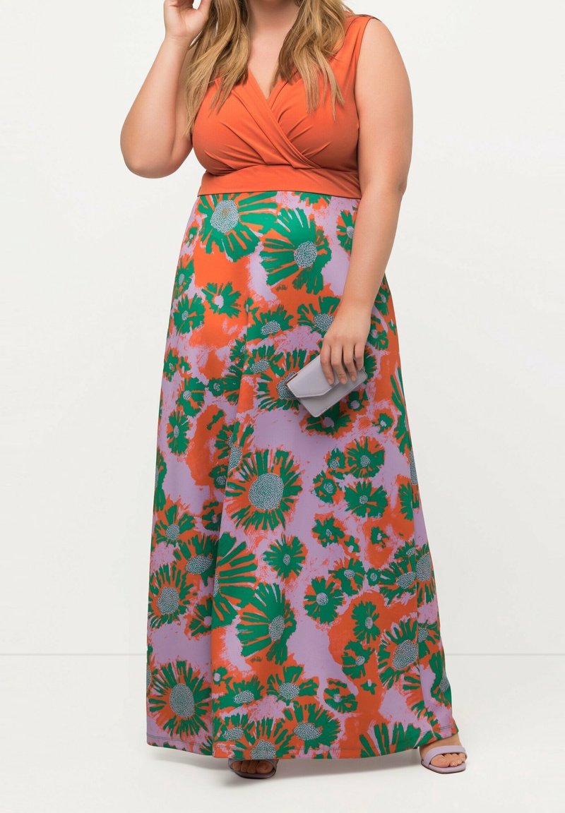 Femme portant un haut sans manches orange et une longue jupe fleurie verte, orange et lavande, tenant une petite pochette grise.