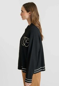 Cardigan nero con accenti ricamati bianchi, chiusura a bottoni, vestibilità ampia, polsini e orlo a coste, mostrato di lato. Materiale misto cotone.