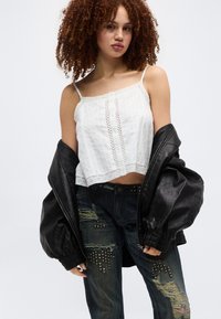 Γυναίκα με σγουρά μαλλιά που φοράει λευκή καμιζόλα με δαντέλα, τζιν με τρυπήματα και τρουκς και μαύρο δερμάτινο μπουφάν σε υπερμεγέθη μέγεθος ριγμένο στους ώμους.