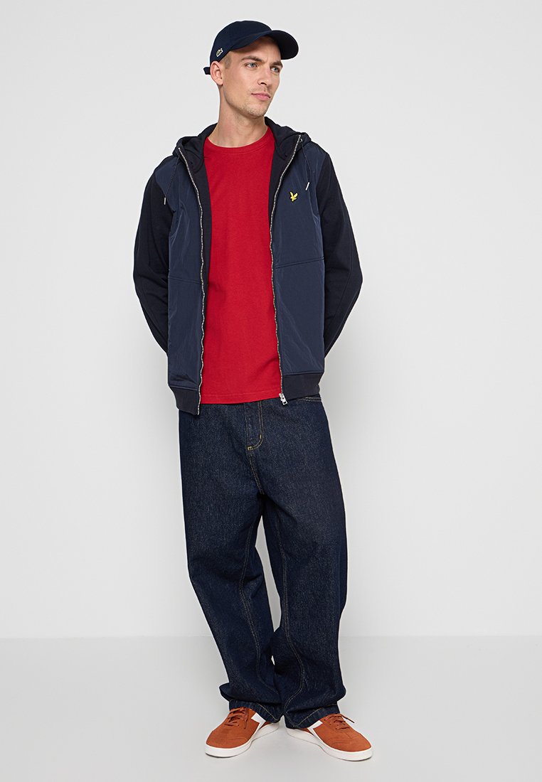 Gilet à capuche navy sur une chemise rouge, associé à un jean en denim sombre ample et des baskets oranges. Le gilet dispose d'une fermeture zippée et de poches avant.