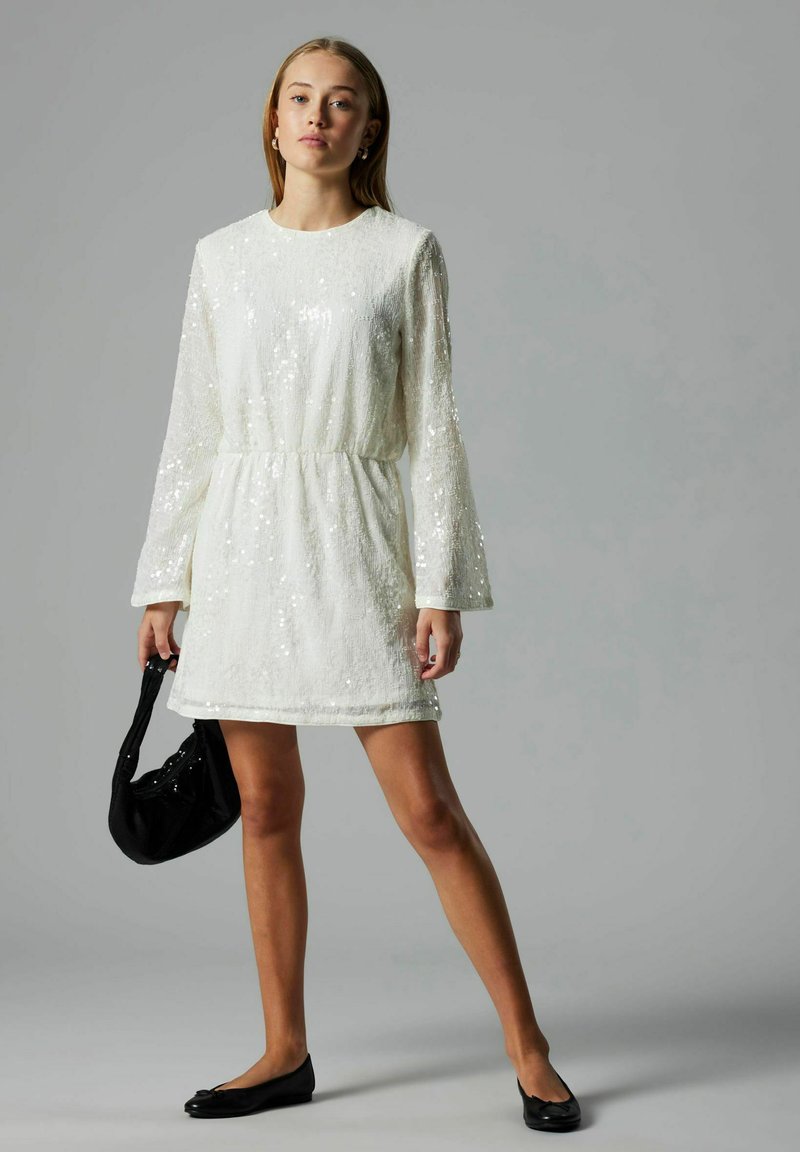 Femme portant une robe mini à manches longues blanche ornée de sequins, tenant un petit sac à main noir et portant des ballerines noires sur un fond gris.