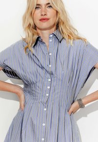 Robe chemise rayée bleue et blanche avec des manches courtes, boutonnière à l'avant et une silhouette large et fluide. Elle présente un tissu texturé et un col classique.