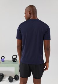 Maglietta atletica navy a maniche corte, tessuto texture e dettagli riflettenti sui lati, indossata con pantaloni corti neri, sullo sfondo ci sono dei manubri.