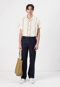 Korte mouwen crèmekleurige blouse met bruine geometrische patronen, marineblauwe broek, witte sneakers en een tan schoudertas. Eenvoudige, ontspannen pasvorm.