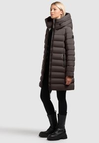 Brązowy puchowy płaszcz z wysokim kołnierzem i kapturem, z pikowanymi sekcjami, zapinanymi bocznymi kieszeniami oraz metalowymi detalami. Noszony z czarnymi legginsami i botkami.