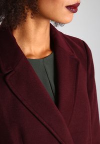 Burgundy ullrock med snedrev, visad över en skoggrön topp; slät textur med rena sömnadsdetaljer.