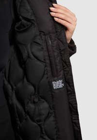 Musta toppatakki tikatulla pinnalla. Siinä on vetoketjullinen tasku ja logoplaatti. Materiaali näyttää sileältä ja kevyeltä.