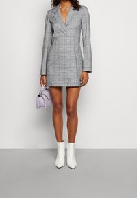 Robe blazer à carreaux gris avec un décolleté en V profond et des manches longues, associée à des bottines blanches et un petit sac à main lavande.