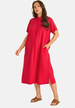 Kobieta ubrana w luźną, czerwoną sukienkę do połowy łydki z krótkimi rękawami i kołnierzykiem, stojąca z jedną ręką w kieszeni i brązowych sandałach.