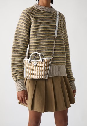 Personne portant un pull rayé beige et gris ainsi qu'une jupe plissée olive, tenant un petit sac à main tissé avec des finitions en cuir blanc et une bandoulière.