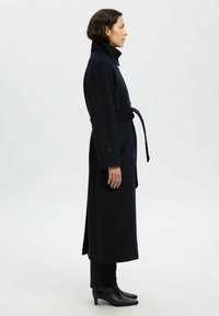 Cappotto lungo in navy realizzato in lana, con colletto alto, vita arricciata, tasche verticali e spacchi laterali. Abbinato a stivali neri con tacco.
