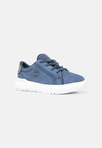 Blue suede sneaker with white rubber sole, tonal stitching, and logo detail. Features laces and a textured heel accent in gray.