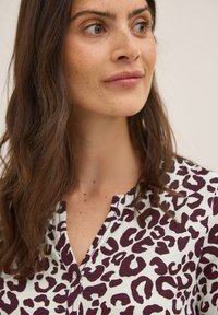 La blusa de estampado de leopardo presenta una base blanca con manchas burdeos profundas y negras, un escote en V y un corte relajado. Tejido suave con una ligera textura.