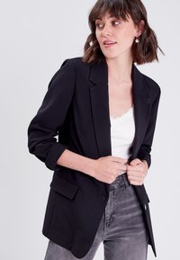 Black blazer with a structured design, single-button closure, and two front pockets. Worn over a white tank top and gray jeans.