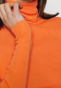 Kvinna som justerar kragen på en ljusorange långärmad polotröja, med handen som rör vid nacken och mörkt hår synligt.