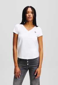 Tricou alb din bumbac cu decolteu în V și mâneci scurte, având un mic logo albastru pe piept, asociat cu blugi gri cu talie înaltă.